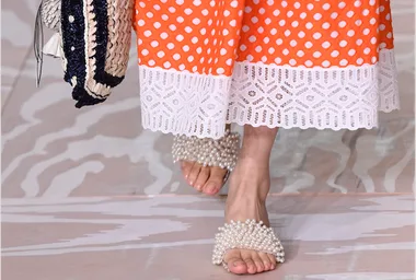 Model wears orange polka dot dress with lace hem and pearl-adorned sandals on the runway.