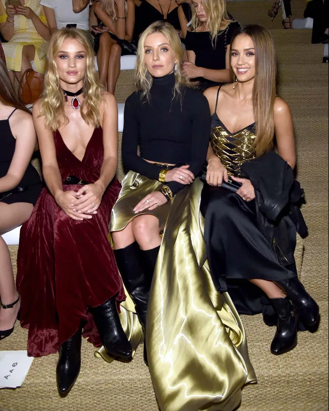 Three women in elegant outfits sitting in a row at a fashion show, with attendees visible in the background.