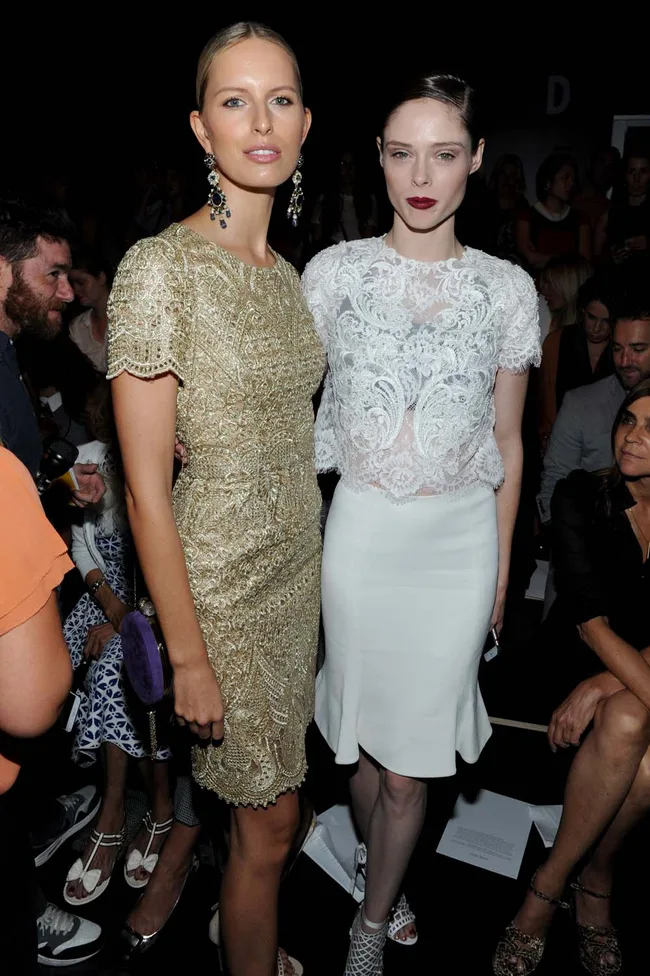 Two women in elegant outfits at a fashion event, one in a gold dress and the other in white lace, surrounded by a crowd.