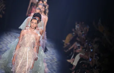 Models in pastel gowns walk the runway, audience taking photos in a dimly lit fashion show venue.