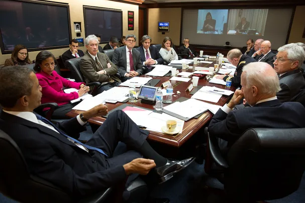 Meeting in a conference room with multiple people around a table, papers and laptops visible, screens on walls.