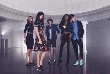Group of five fashionably dressed models posing in a large, bright room with high ceilings and columns.