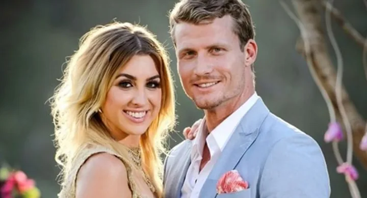A man in a light blue suit and a woman in a gold dress smiling and posing outdoors with trees in the background.