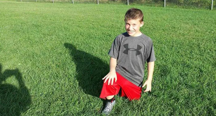 A boy kneels on grass wearing a gray Under Armour shirt and red shorts, smiling in the sunshine.