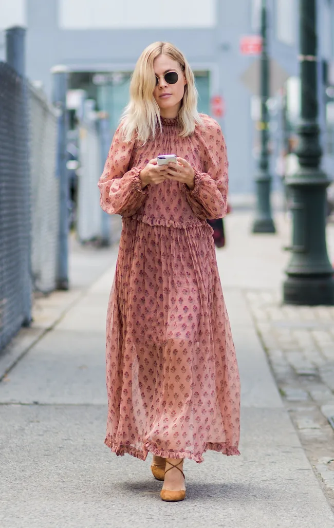 Woman in a pink floral dress and sunglasses walking on a city sidewalk, holding a smartphone.
