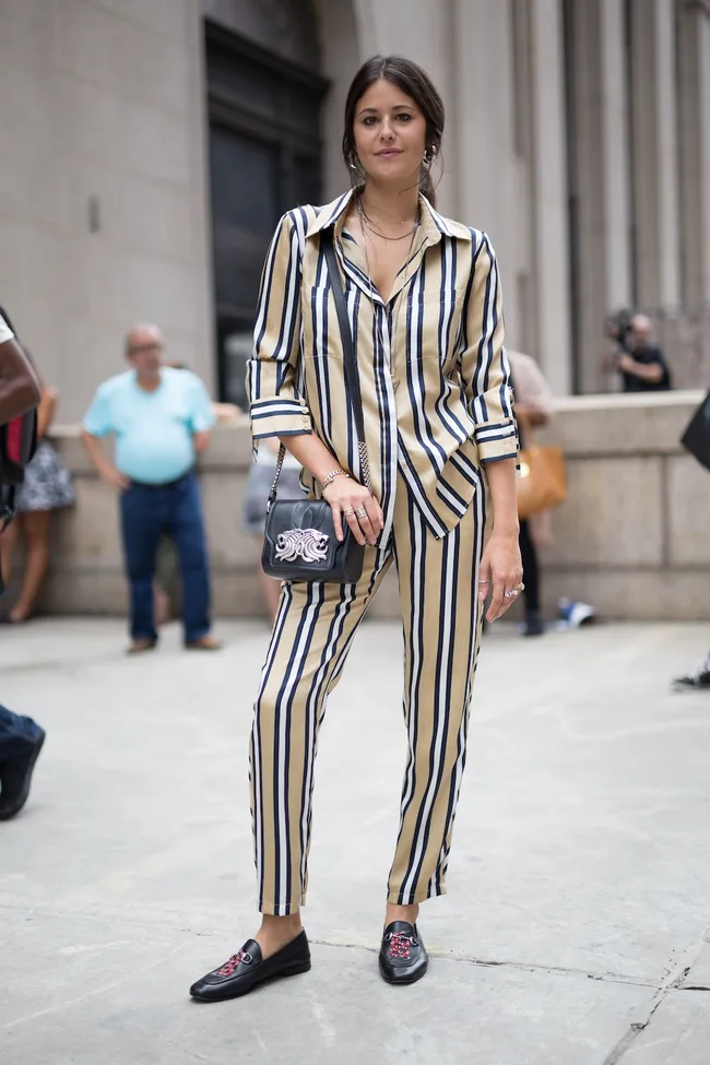 Woman in striped outfit with matching top and trousers, posing outdoors with a small black handbag and loafers.