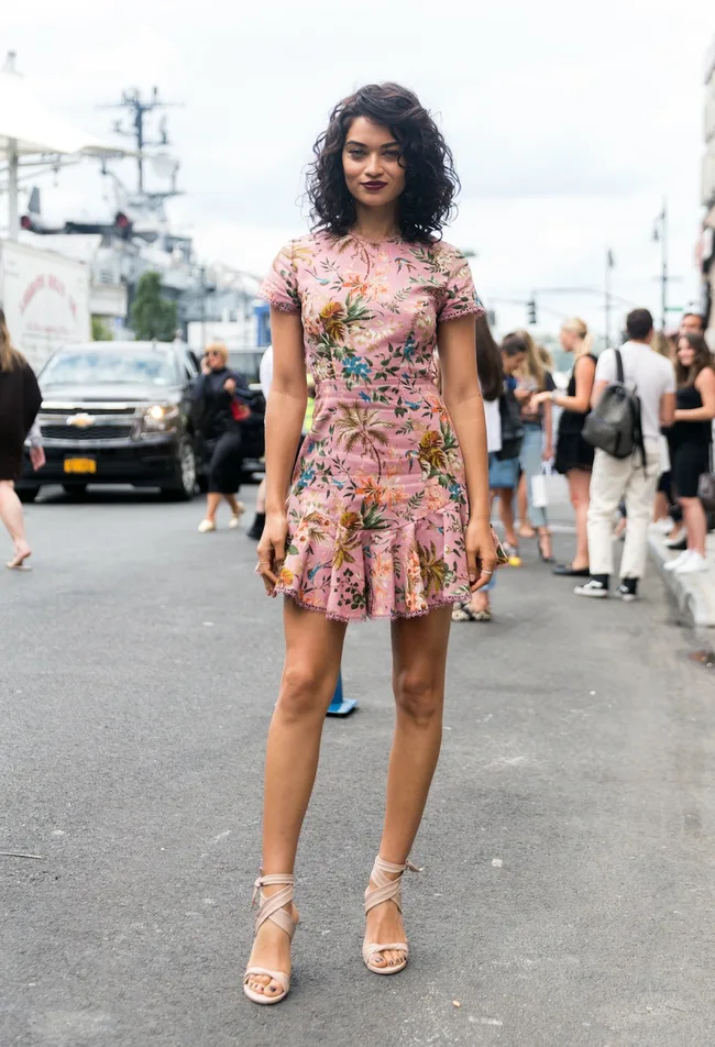 Woman in a pink floral dress and heels stands on a city street, with a group of people in the background.