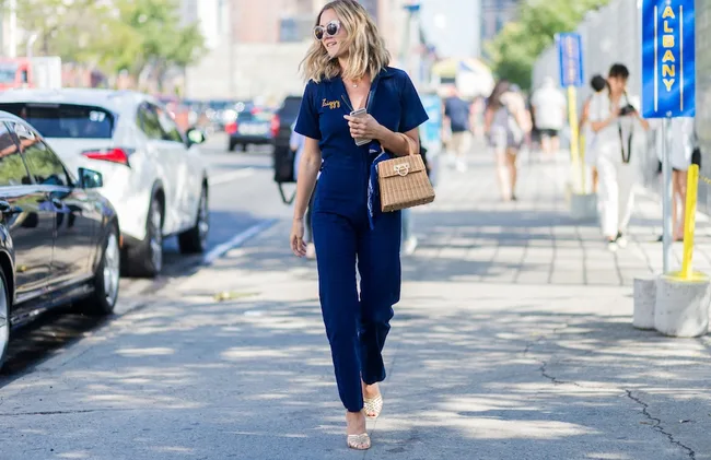 Woman in blue jumpsuit walking on a city sidewalk, carrying a brown woven handbag and wearing sunglasses.