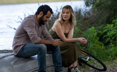 A man and woman sit on a boat's edge by a lakeside, talking with a bicycle in the background.