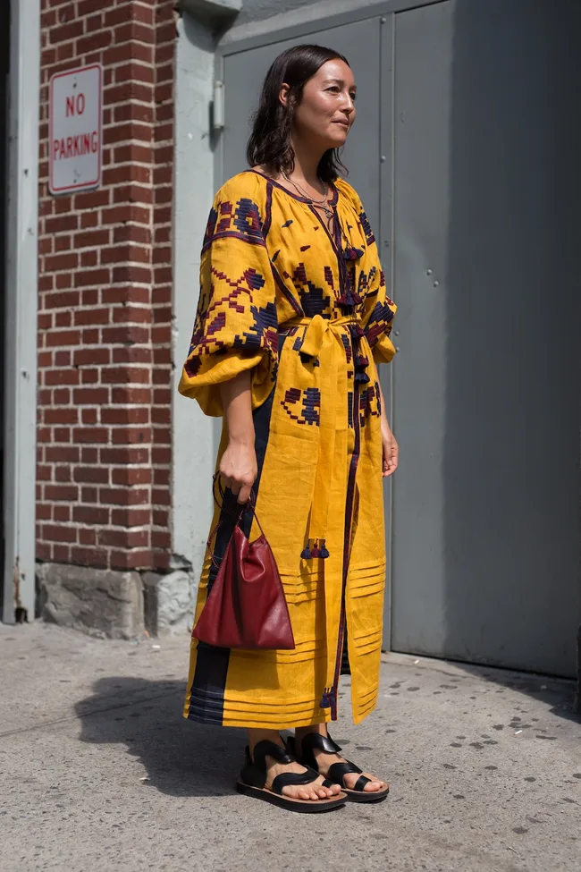 Woman in a yellow patterned dress and black sandals, holding a red bag, stands outside near a "No Parking" sign.