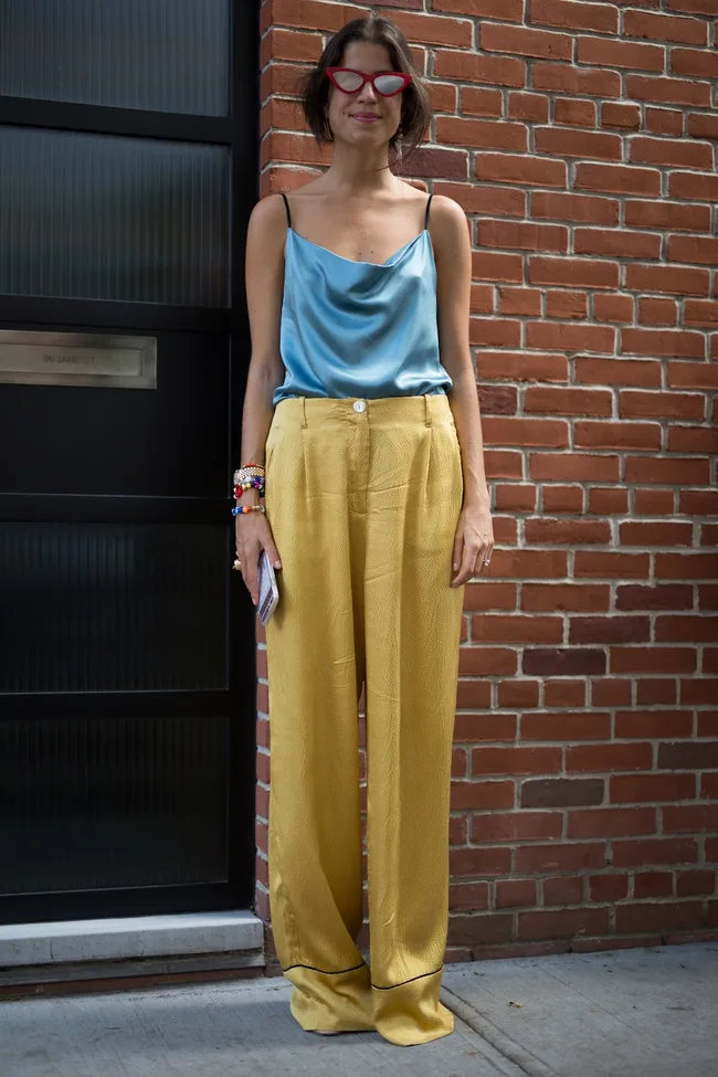 Person in blue top, yellow pants, red cat-eye glasses, holding a phone, standing against a brick wall.