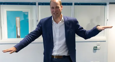 A man in a blue suit with outstretched arms stands in a room with white walls and windows.