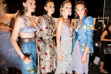 Models in colorful floral outfits backstage at a fashion show, wearing statement earrings.
