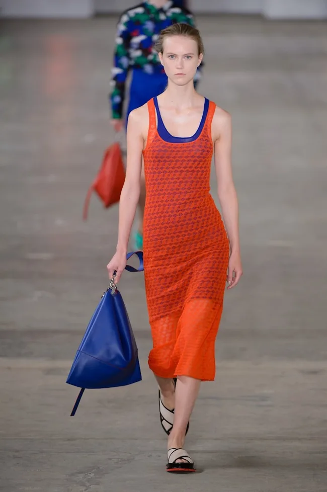 Model in an orange and blue dress, holding a blue bag, walks the runway in a fashion show.