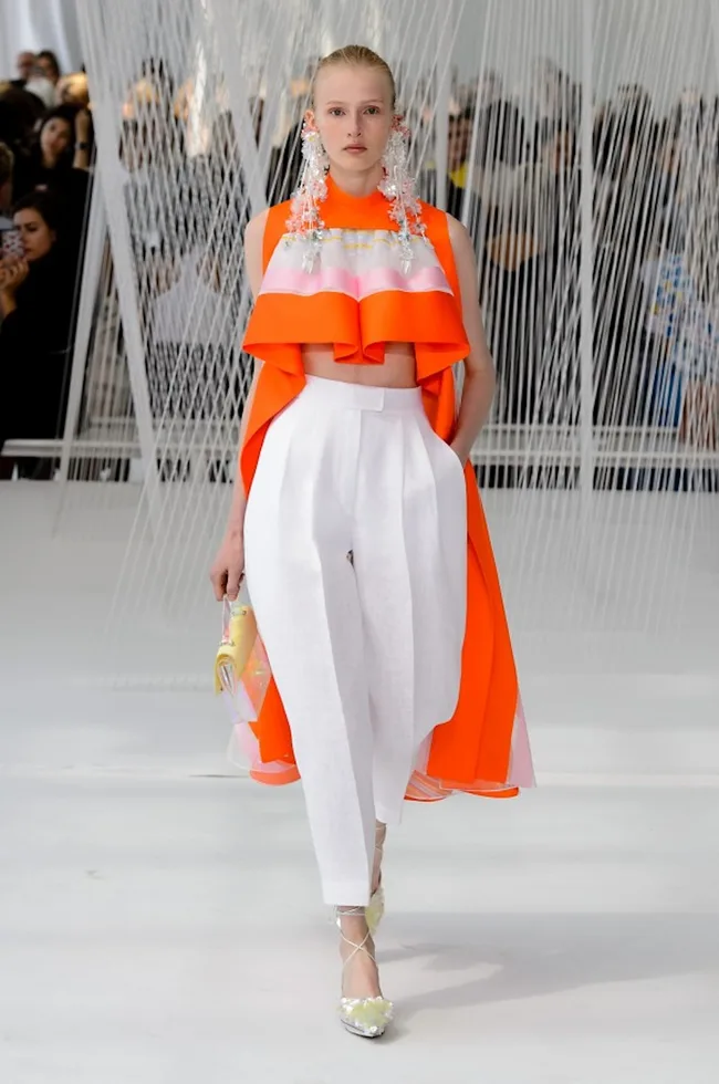 Model in an orange top with pink accents and white pants, wearing large earrings, walking on a runway.