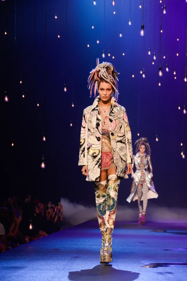 Model walking runway in colorful, embroidered outfit and boots, with dramatic braided hairstyle, under hanging lights.