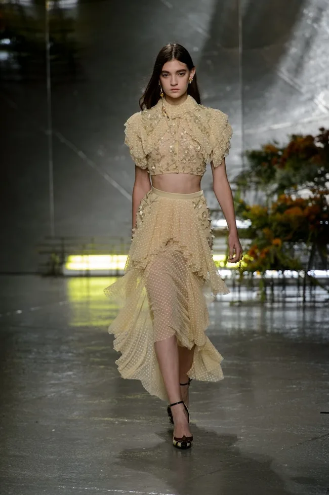 Model in a beige floral crop top and layered skirt walks on a reflective runway with blurred floral background.