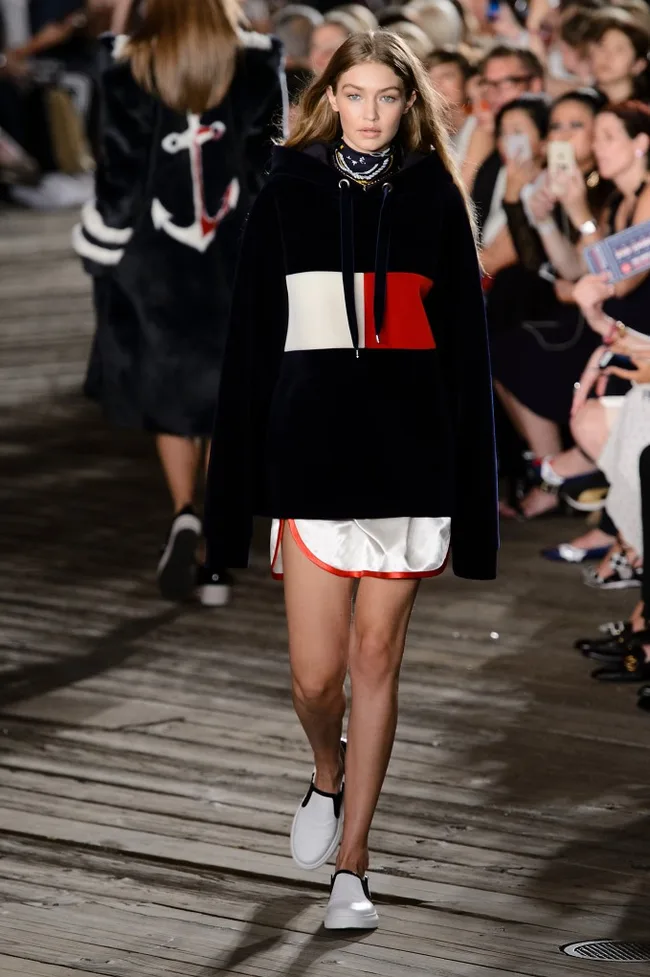Model walking the runway in a chic black, white, and red outfit at a fashion show, surrounded by a seated audience.