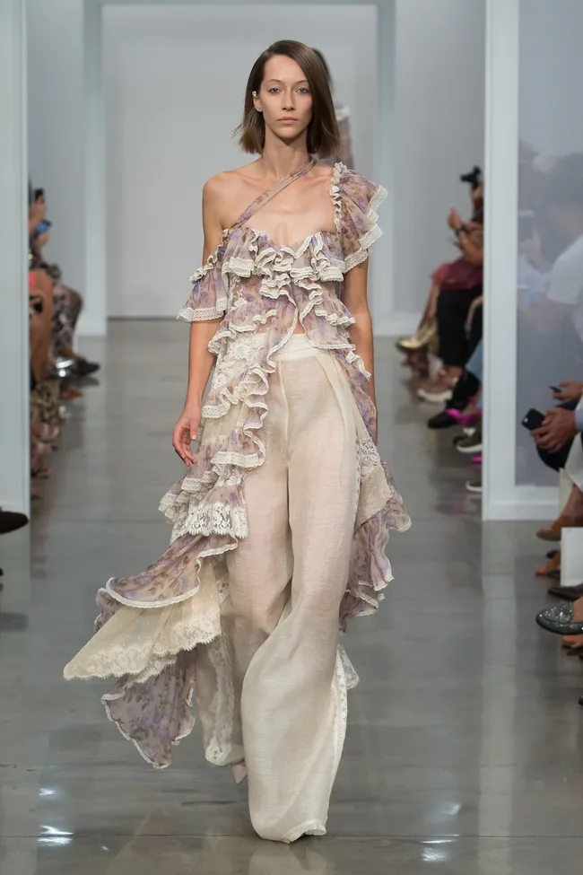 Model wearing a ruffled, asymmetrical floral dress on the runway, surrounded by seated spectators.