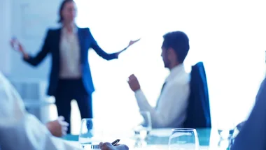 A person giving a presentation at a business meeting with a blurred background of seated individuals and a table with glasses.