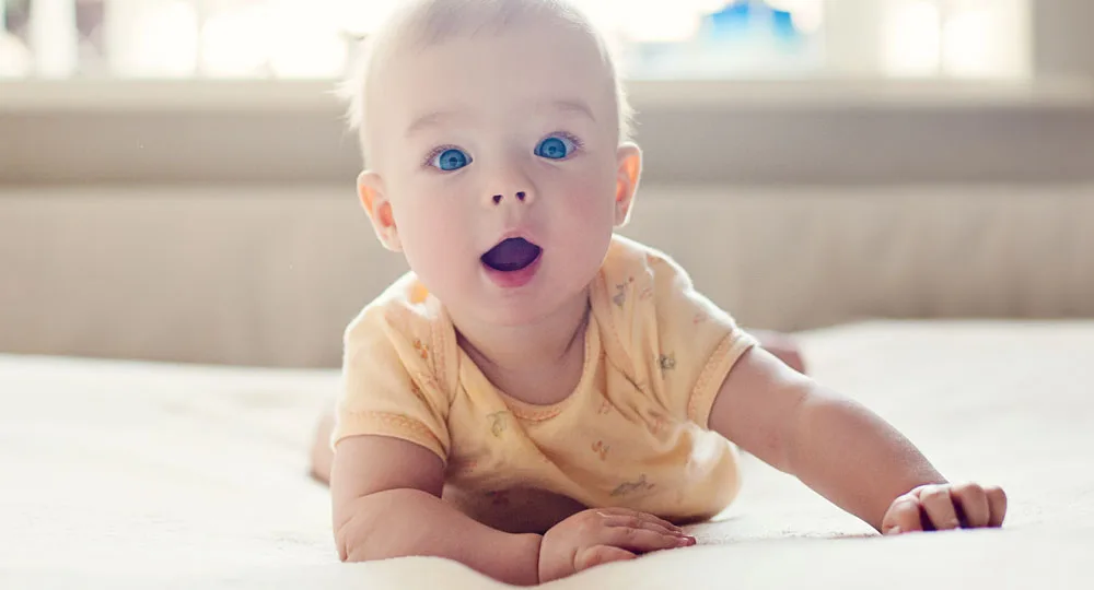 A baby with blue eyes, wearing a yellow onesie, lies on a bed with an expression of curiosity and surprise.