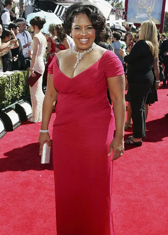 Elegant woman in a red gown, smiling on a red carpet at an outdoor event, surrounded by people and cameras.