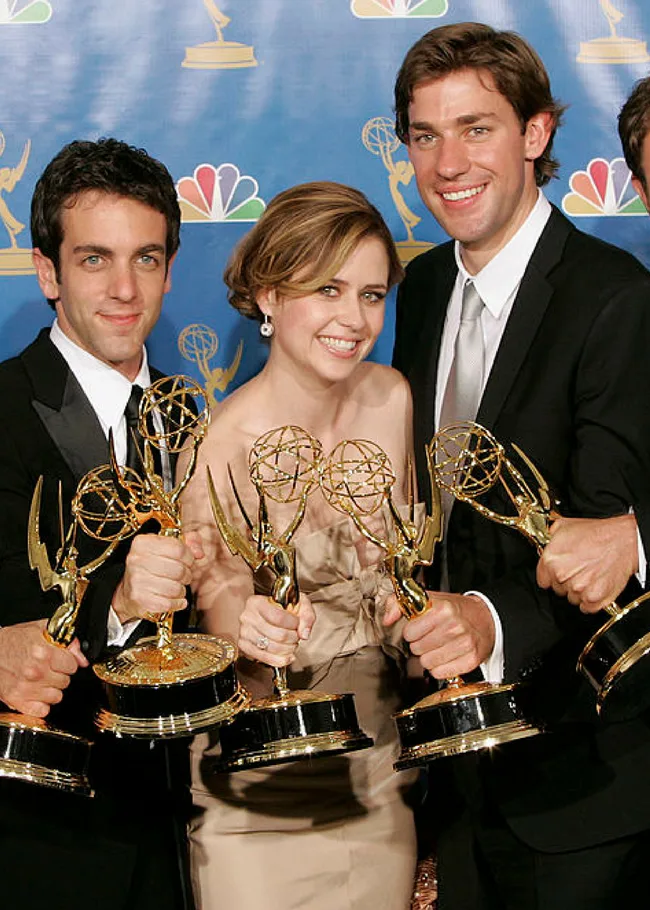 Three people smiling and holding Emmy Awards in front of a blue background with Emmy logos and the NBC logo.