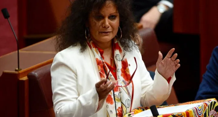 A woman in a white blazer speaks at a podium with colorful artwork, holding glasses in a legislative chamber.