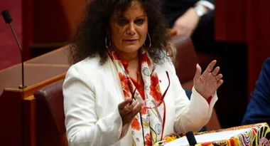 A woman in a white blazer speaks at a podium with colorful artwork, holding glasses in a legislative chamber.