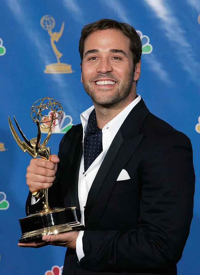 Actor smiling while holding an Emmy award against a blue background with NBC logos.