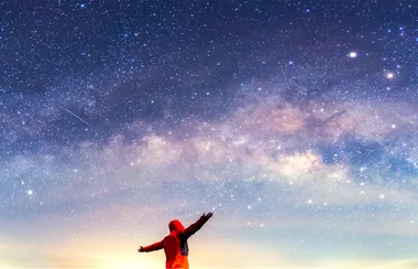 Person in red jacket with arms raised, gazing at a starry night sky with Milky Way visible.