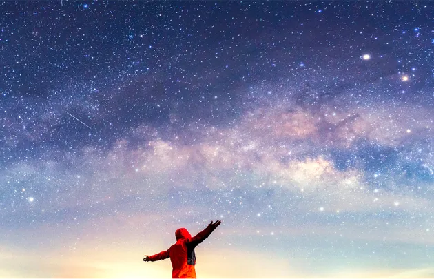 Person in red jacket with arms raised, gazing at a starry night sky with Milky Way visible.