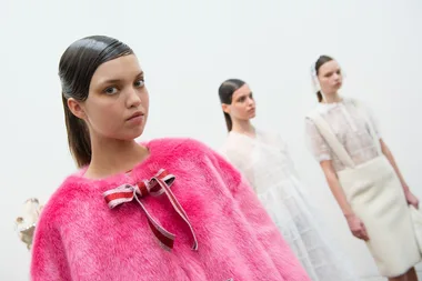 Models wearing high fashion outfits; one in a vibrant pink fuzzy top with a bow in focus, others in white, blurred background.