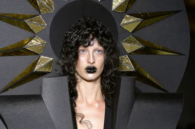 Model with curly hair, black lipstick, wearing avant-garde geometric outfit, gold and black starburst backdrop at fashion show.