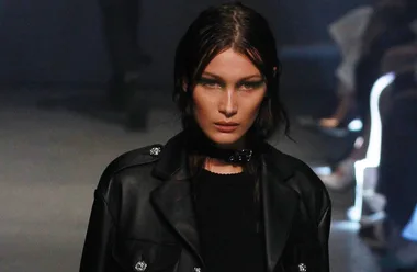 A model wearing a black leather coat walks on a dimly lit runway.