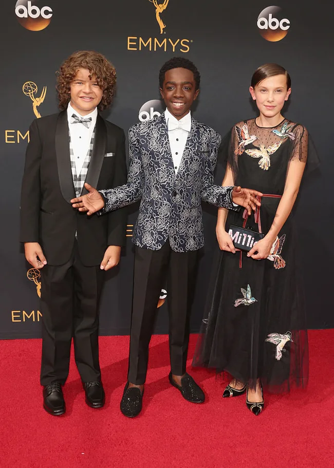 Three young actors in formal attire pose on the Emmys red carpet.