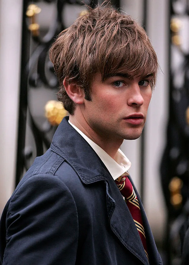 Young man with brown hair in a dark coat and striped tie, looking over his shoulder, in front of a decorative iron gate.