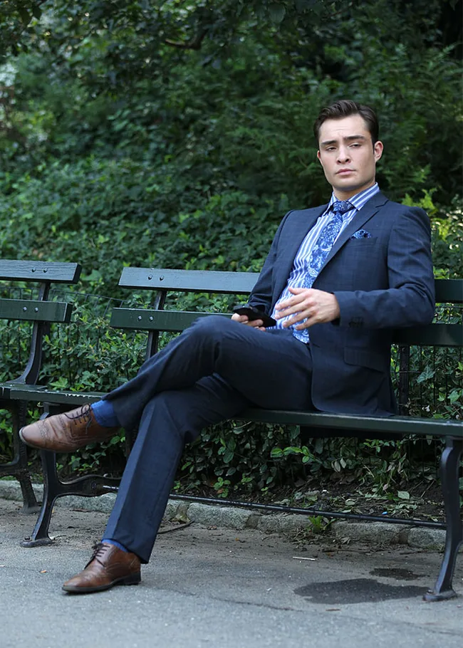 Man in a blue suit sitting on a park bench, holding a phone, with a lush green background.