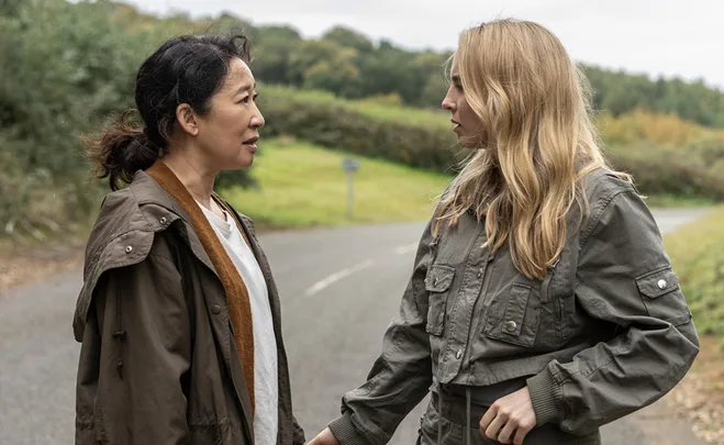 Two women face each other on a rural road, engaged in conversation, wearing jackets, with green landscapes in the background.