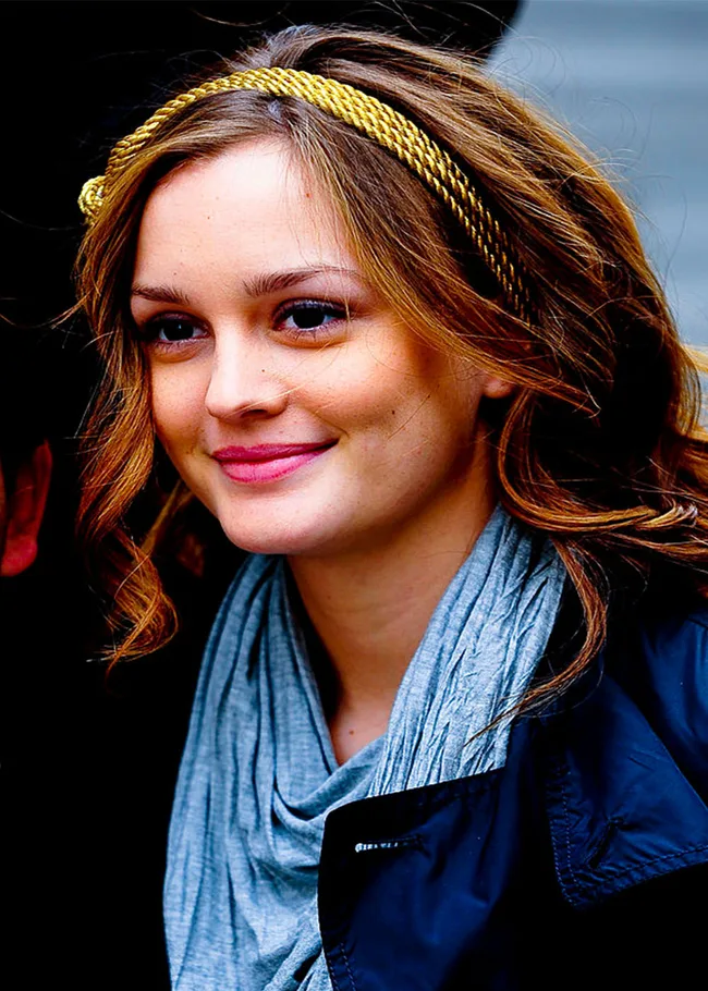 Young woman with wavy hair and a gold headband, smiling in a blue scarf and dark jacket.