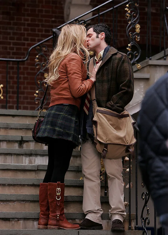 A man and woman kiss on outdoor stairs, with the woman in a brown jacket and boots, and the man holding a messenger bag.