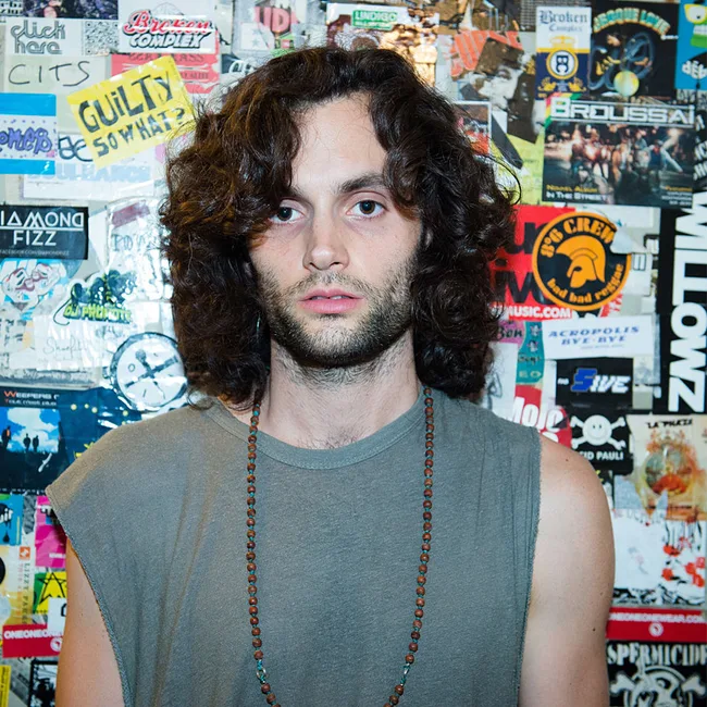 Man with long curly hair and a beaded necklace stands in front of a wall covered in posters and stickers.