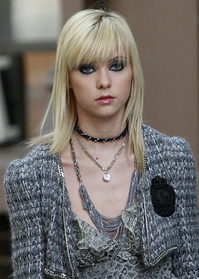 Blonde woman with smoky eye makeup, wearing layered necklaces and a gray tweed jacket, gazing forward.