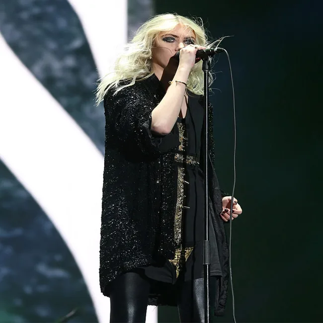 Singer in black outfit performing on stage with microphone under colorful lights.