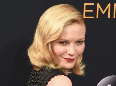 A person with blonde hair poses on a red carpet, wearing a black textured dress, with a confident expression.