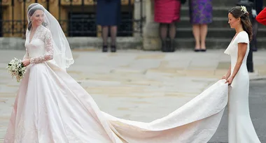 Bride in a lace wedding dress holds bouquet, while another woman carries her train outside a building.