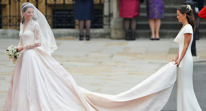 Bride in a lace wedding dress holds bouquet, while another woman carries her train outside a building.