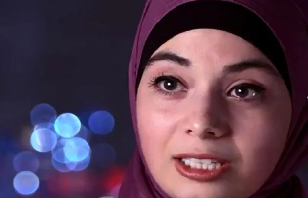 A woman wearing a purple hijab is speaking with blurry blue and purple lights in the background.