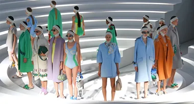 Models on tiered steps wearing retro swimwear and coats in pastel colors, accessorized with headbands and sunglasses.