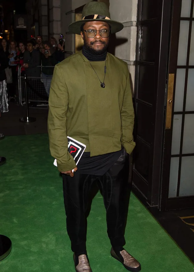 A person in green jacket and hat stands on a green carpet, holding a tablet, with a crowd in the background.